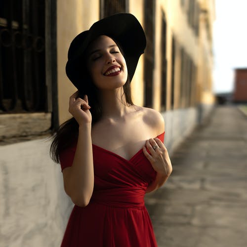 看着上面和闭上眼睛时，按住黑色太阳帽的红色露肩连衣裙的女人 · 免费素材图片