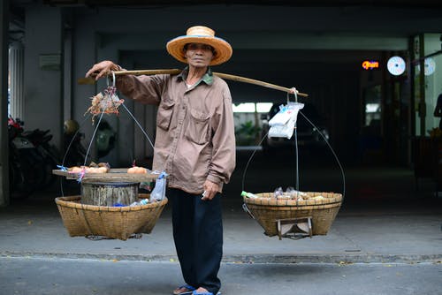 棕色夹克的男人，两个篮子吊在肩膀上的木杆 · 免费素材图片