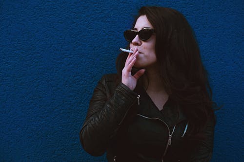 女人站在蓝色的墙壁上抽着烟的照片 · 免费素材图片