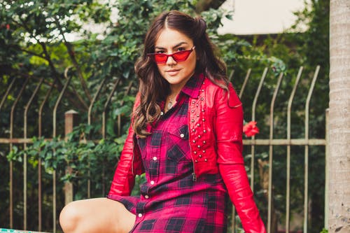 坐在灰色金属栅栏前的红色皮夹克和太阳镜的女人的照片 · 免费素材图片