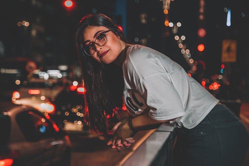 夜间在人行道上斜倚在扶手上的眼镜的微笑妇女的照片 · 免费素材图片