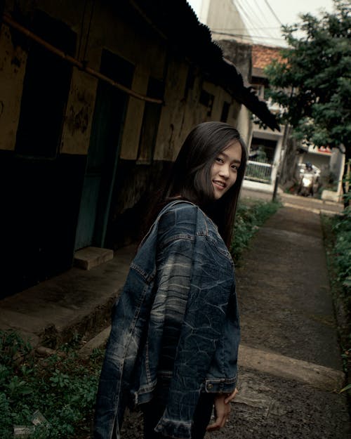 站在一条小巷，肩上背着蓝色牛仔夹克的微笑妇女的照片 · 免费素材图片