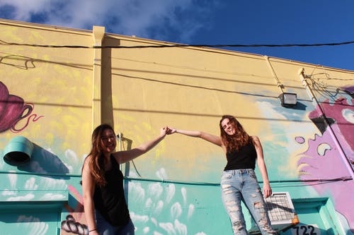 两个笑女人站在涂鸦墙附近时握着的手的照片 · 免费素材图片
