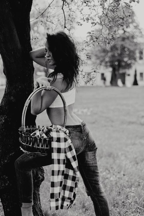女人靠在树上摆姿势时提着篮子的灰度照片 · 免费素材图片
