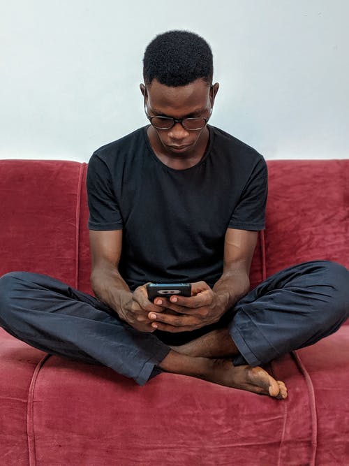 男子坐在红色绒面革沙发上，拿着黑色的android智能手机 · 免费素材图片