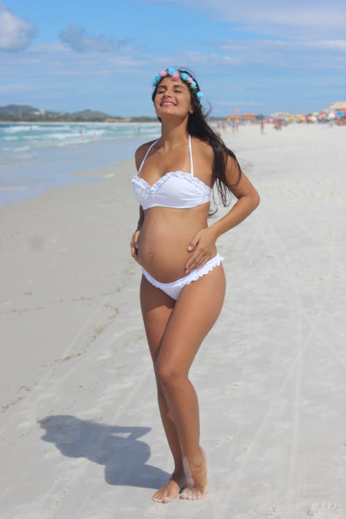 孕妇在白色比基尼泳装站立和微笑在海边，闭着眼睛的照片 · 免费素材图片