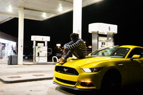 坐在黄色的福特野马引擎盖上的人的照片停在加油站 · 免费素材图片