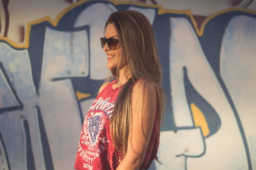 红色背心和太阳镜涂鸦墙附近的微笑妇女的侧视图照片 · 免费素材图片