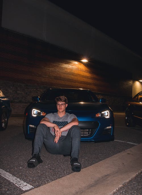 男人穿着灰色圆领衬衫和灰色运动裤坐在蓝色的车辆前 · 免费素材图片