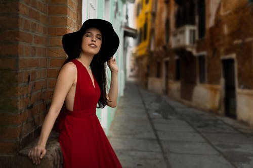 闭着眼睛，穿着一件红色的连衣裙和黑色的帽子，斜倚在胡同中的红砖墙上的女人的照片 · 免费素材图片
