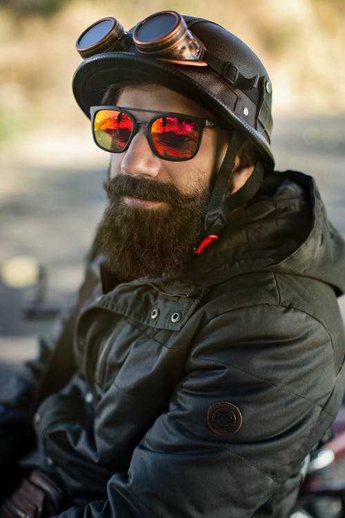 胡子的男子戴着黑色摩托车头盔与黄铜护目镜，太阳镜和黑色连帽外套的特写照片 · 免费素材图片