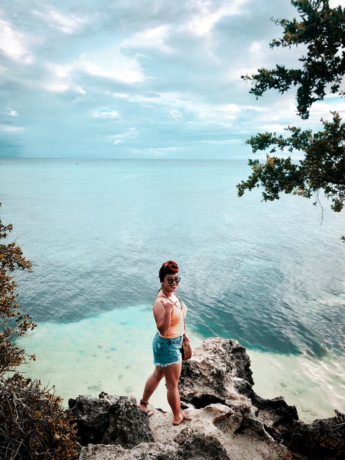 站在海边岩石在海边附近的蓝色牛仔短裤的微笑女人的照片 · 免费素材图片