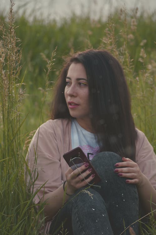 女人拿着手机坐在草地上的照片 · 免费素材图片
