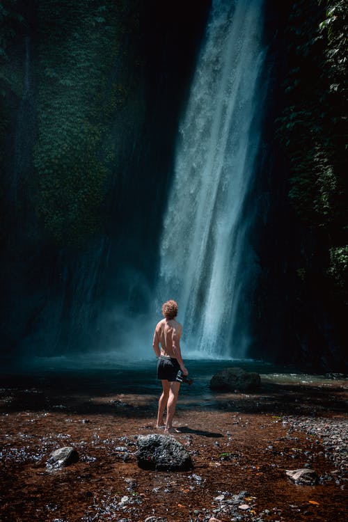 男子站在瀑布附近的岩石上的照片 · 免费素材图片