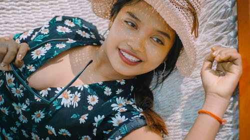 微笑的女人，在黑色的花顶和持有太阳镜躺在白色的吊床上，看着别处的太阳帽的特写照片 · 免费素材图片