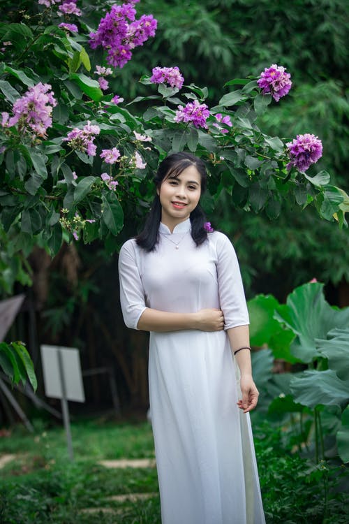站在紫色的花下的白色连衣裙的微笑女人的照片 · 免费素材图片