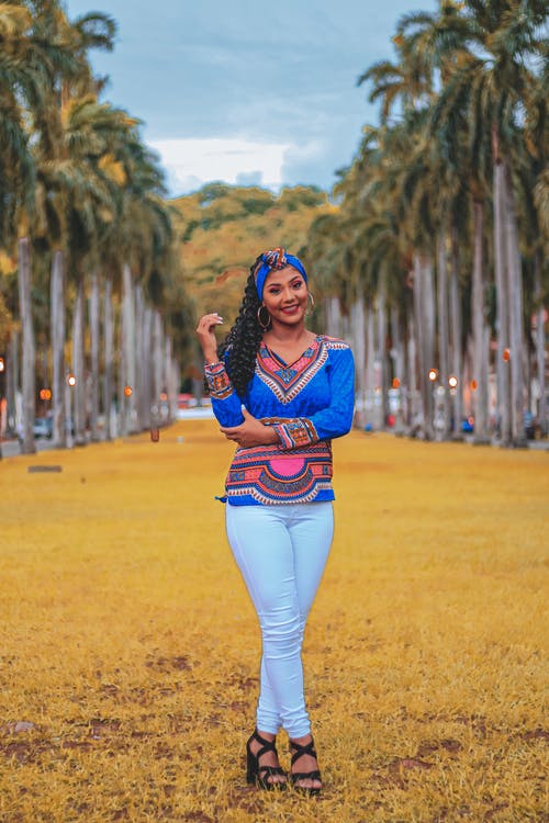 微笑的女人，穿着蓝色dashiki和白色的牛仔裤，站在黄色的草丛中，在棕榈树旁摆姿势的照片 · 免费素材图片