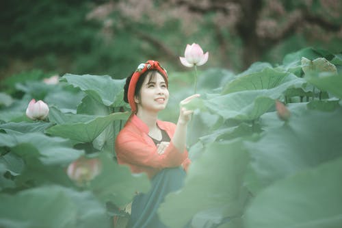 坐在花田中间时拿着一朵粉红色的花的微笑妇女的照片 · 免费素材图片