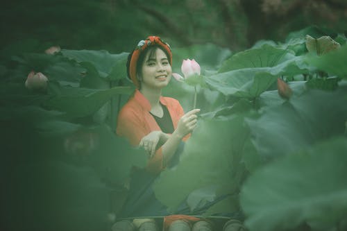 坐在花田中间时拿着一朵粉红色的花的微笑妇女的照片 · 免费素材图片