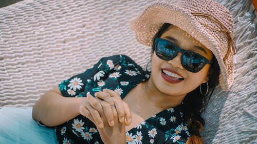 微笑的女人，在黑色的花顶，太阳帽和躺在白色的吊床上的太阳镜的特写照片 · 免费素材图片