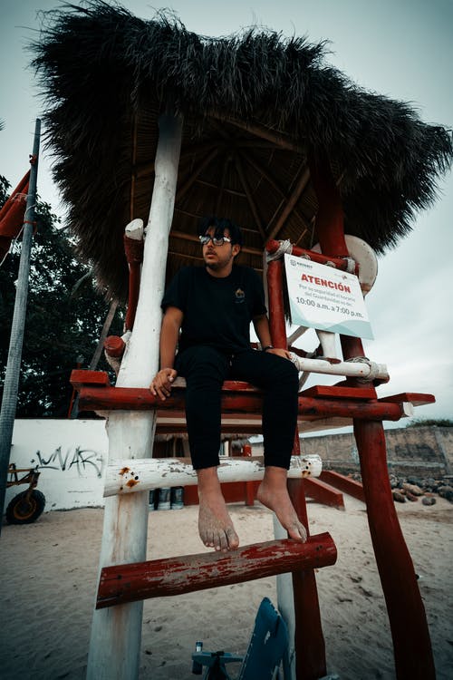 穿着黑色t恤和裤子坐在救生塔中的赤脚男人的照片 · 免费素材图片