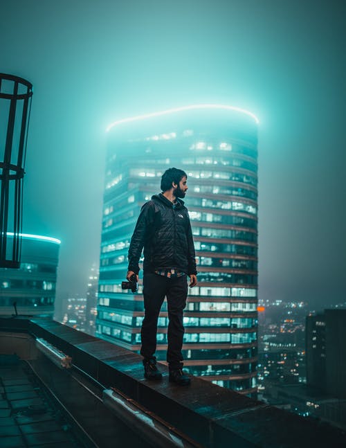男子手持单反相机走在俯瞰夜晚的建筑物边缘的照片 · 免费素材图片