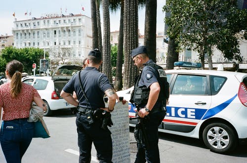 两名警察站在路上白色和蓝色警车旁边另一个人附近 · 免费素材图片