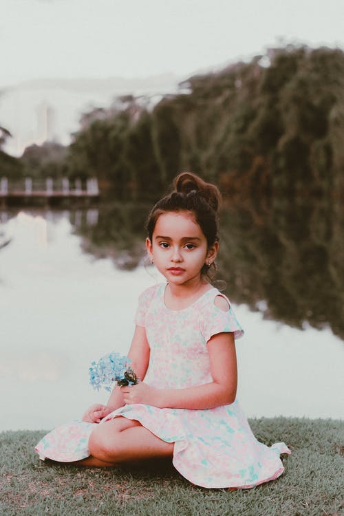 年轻的女孩穿着白色花裙子的化妆，坐在水边的草地上，同时手捧鲜花的照片 · 免费素材图片