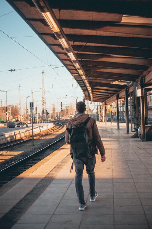 后视图的人，穿着棕色连帽衫，灰色的裤子和背包独自走在空火车站站台 · 免费素材图片