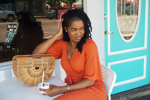 坐在店外拿着冰淇淋，同时看着别处的橙色v领短袖连衣裙的微笑女人的照片 · 免费素材图片