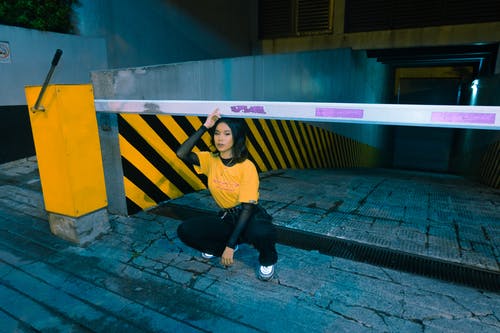 黄色的t恤和黑色的裤子蹲在栅栏门附近的女人的照片 · 免费素材图片