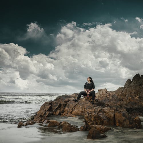 女人坐在岩石附近海滩望着远方的照片 · 免费素材图片
