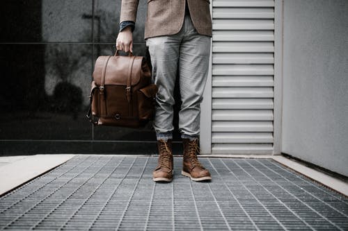 棕色西装外套，灰色的裤子和棕色的靴子，拿着棕色的皮包，站在建筑物外面的人的照片 · 免费素材图片