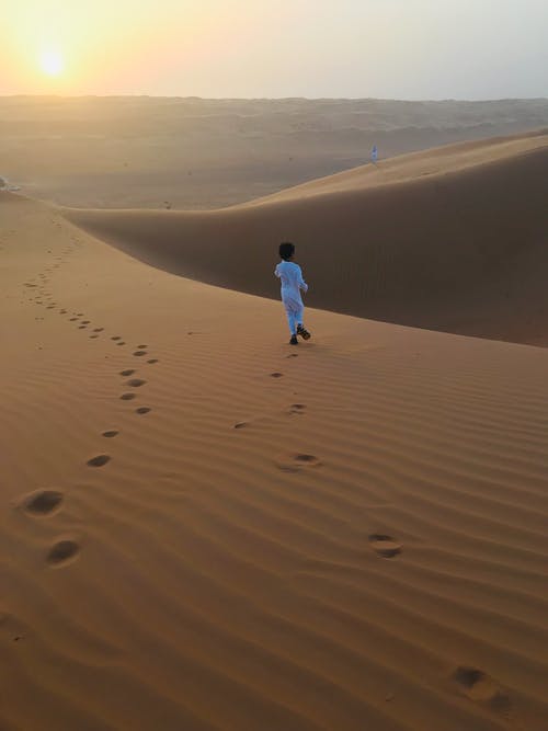 在金色小时内独自走在沙漠沙地上的白色thobe的穆斯林男孩的后视图照片 · 免费素材图片