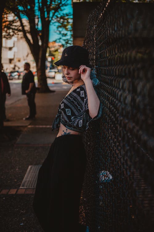 黑色礼服顶部，帽子和底部倾斜在链节围栏上的人行道上摆姿势的女人的照片 · 免费素材图片