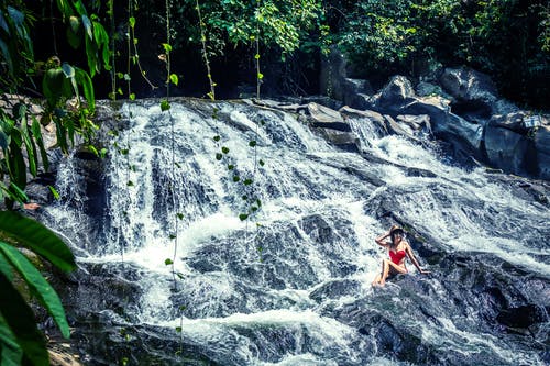 女人坐在瀑布附近的岩石上的照片 · 免费素材图片