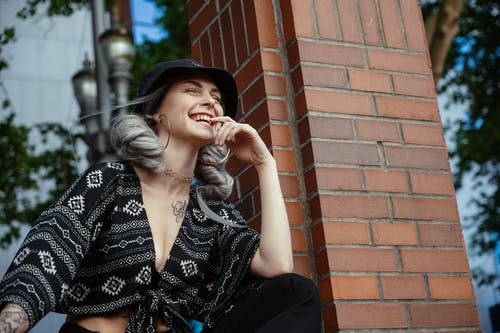 黑顶摆姿势，她的眼睛在砖支柱旁边闭着笑的女人的照片 · 免费素材图片