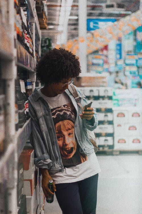 倾斜在超市货架上携带电话时携带玻璃瓶的女人的照片 · 免费素材图片
