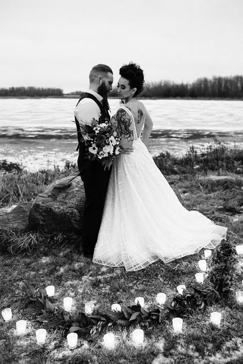 男人和女人穿着婚纱站在灰色的照片中的柱子蜡烛包围的水体附近 · 免费素材图片