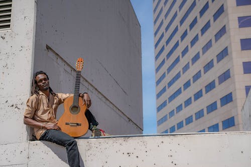 一个男人穿着棕色衬衫，坐在混凝土板上拿着吉他的选择性聚焦摄影 · 免费素材图片