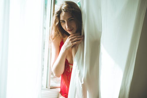 站在白色的窗帘后面的红色内衣的微笑妇女的照片 · 免费素材图片