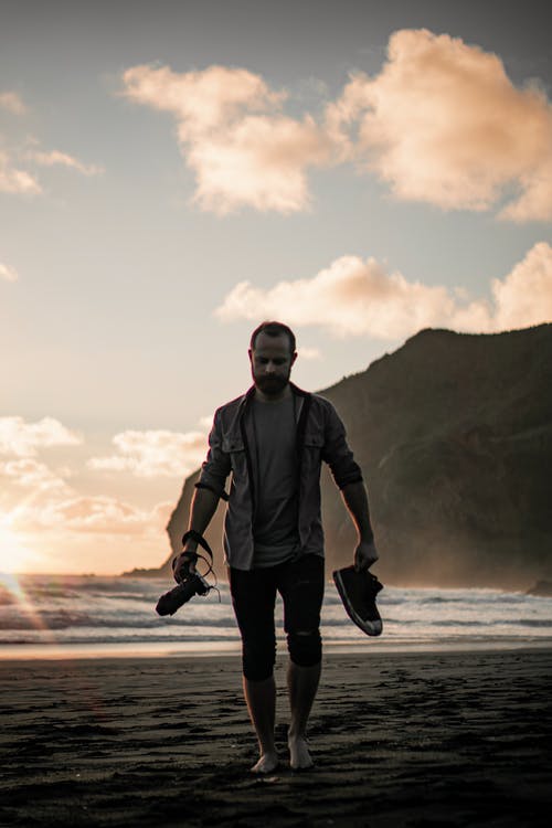男子拿着鞋子和相机走在沙滩上 · 免费素材图片