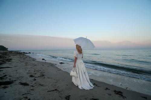 穿着白色连衣裙的女人背着伞，独自一人走在沙滩上时背着手提包的背影照片 · 免费素材图片