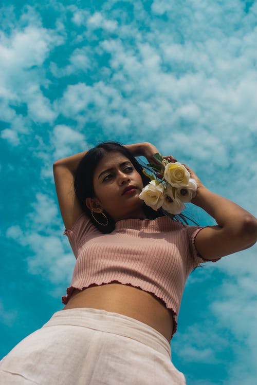 女人在桃短袖作物顶部持有白玫瑰摆在蓝蓝的天空下的低角度照片 · 免费素材图片
