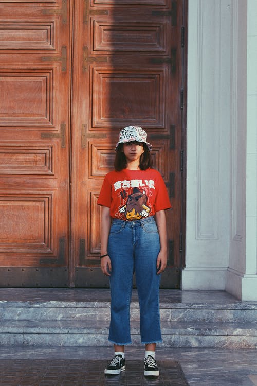 十几岁的女孩，穿着水桶帽子，红色t恤和蓝色牛仔裤站在棕色木门前的照片 · 免费素材图片
