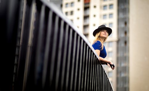 戴着蓝色上衣和帽子靠在金属栏杆上的女人 · 免费素材图片