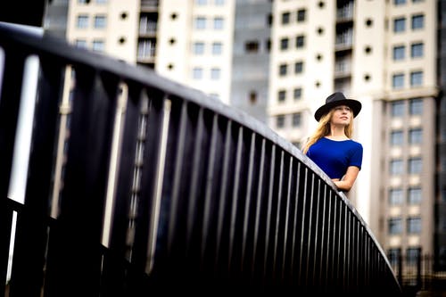 女人穿着蓝色上衣和一顶帽子，倚在金属栏杆 · 免费素材图片
