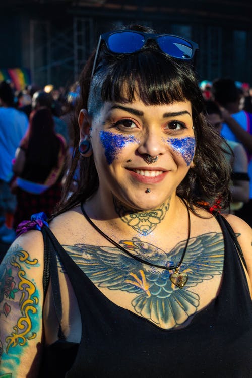 微笑的纹身的女人在黑色背心和太阳镜，蓝色的闪光在她的脸颊与人群中合影的背景上的肖像照片 · 免费素材图片
