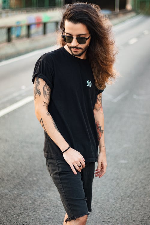纹身男子穿着黑色t恤，太阳镜和黑色牛仔短裤摆在马路中间而俯视的照片 · 免费素材图片