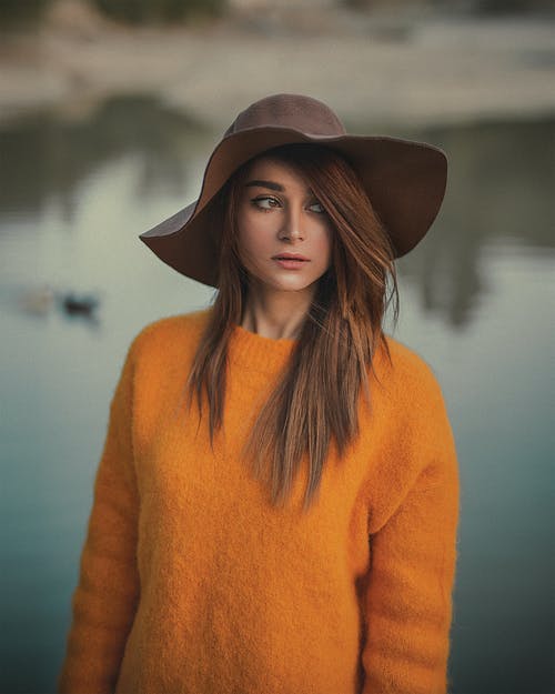 女人穿着橙色毛衣和棕色帽子摆在背景中的水面看着别处的照片 · 免费素材图片